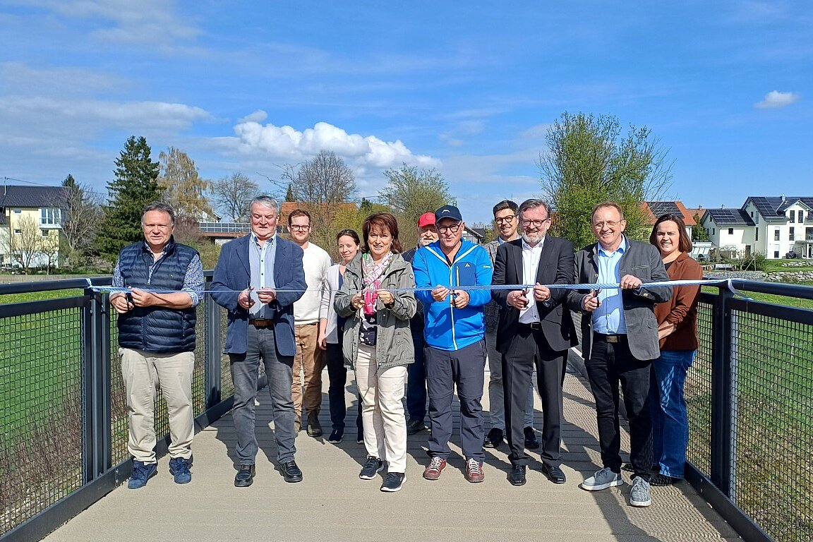Eine Gruppe von Personen steht auf einer Brücke und hält ein Band, das sie gleich durchschneiden wollen. Im Hintergrund sind Bäume und Häuser sichtbar. Es ist ein sonniger Tag mit blauem Himmel.