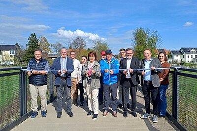 Eine Gruppe von Personen steht auf einer Brücke und hält ein Band, das sie gleich durchschneiden wollen. Im Hintergrund sind Bäume und Häuser sichtbar. Es ist ein sonniger Tag mit blauem Himmel.