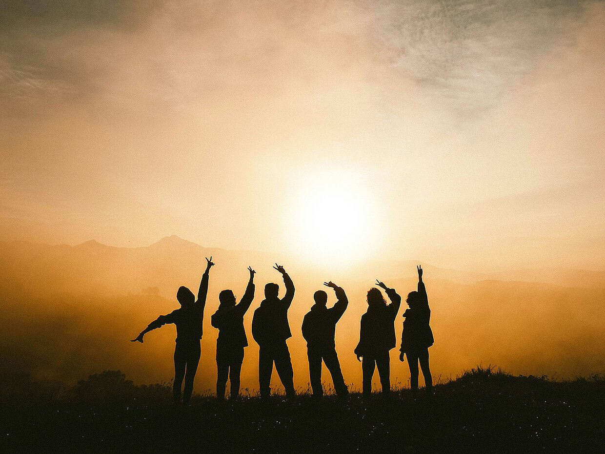 Silhouette von sechs Personen, die vor einem Sonnenuntergang stehen. Sie heben die Arme in die Luft und erscheinen vor einem nebligen Hintergrund. Die Landschaft ist sanft hügelig und von warmen Farben durchzogen. Die Stimmung wirkt fröhlich und enthusiastisch.