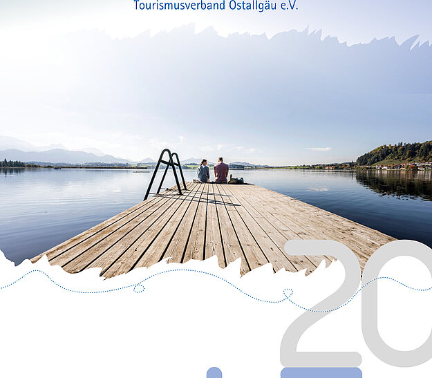 © Tourismusverband Ostallgäu e.V. Ein ruhiger See mit einer Holzplattform, auf der zwei Personen sitzen. Über dem Wasser schimmert der Himmel in sanften Blau- und Grautönen. Der Titel "Jahresbericht" und der Untertitel "Tourismusverband Ostallgäu e.V." sind prominent oben platziert.