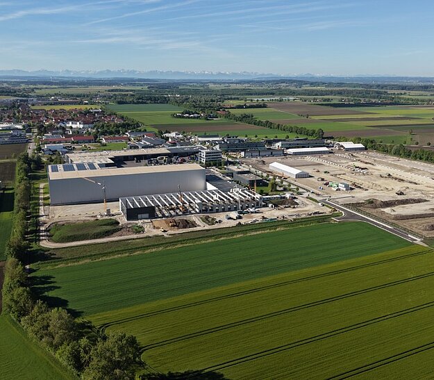 Luftaufnahme eines Industriegebiets mit mehreren großen Gebäuden und Lagerflächen, umgeben von grünen Feldern und landwirtschaftlichen Flächen. Im Hintergrund sind die Konturen von Städten und Bergen zu sehen. Die Landschaft ist weitläufig und gut strukturiert.