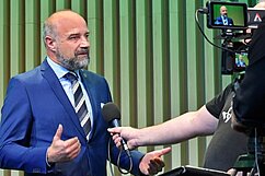 Ein Mann mit Bart und Anzug steht vor einer Kamera und spricht mit einem Interviewer. Der Hintergrund ist in sanften Grüntönen gehalten. Der Mann gestikuliert während des Gesprächs.