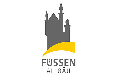 Logo von Füssen im Allgäu, das eine stilisierte graue Burg mit Türmen zeigt. Darunter steht der Schriftzug „Füssen“ in Großbuchstaben und „Allgäu“ in Kleinbuchstaben. Eine gelbe, geschwungene Linie ergänzt das Design.