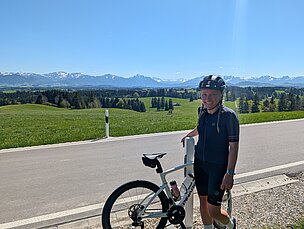 Eine Person steht neben einem Fahrrad auf einer Straße, umgeben von grünen Wiesen und Bergen im Hintergrund. Der Himmel ist klar und blau, und die schneebedeckten Gipfel der Berge sind sichtbar. Die Person trägt einen Helm und sportliche Kleidung.