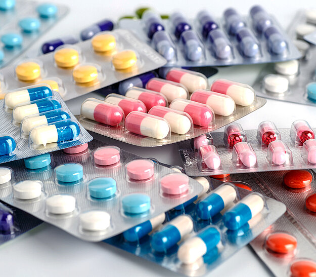 Eine Vielzahl von Blisterverpackungen mit verschiedenen Arten von Tabletten und Kapseln in unterschiedlichen Farben, darunter gelbe, blaue, rosa und rote Pillen, ist chaotisch auf einer Oberfläche angeordnet.