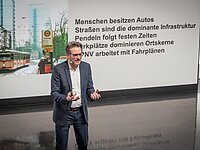 Ein Mann in einem Anzug steht auf einer Bühne und spricht. Im Hintergrund ist eine Präsentation mit Text zu sehen, der Themen wie Autoverkehr, Straßeninfrastruktur und Nahverkehr behandelt. Der Raum ist modern gestaltet.