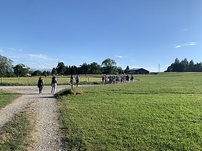 Eine Gruppe von Menschen wandert auf einem Schotterweg durch ein weites, grünes Feld unter blauem Himmel. Im Hintergrund sind Bäume und Gebäude zu sehen.