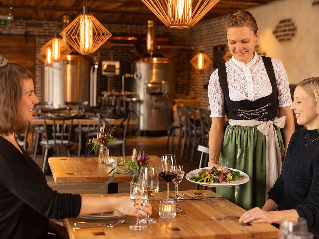 Eine Kellnerin in traditioneller Kleidung serviert ein Gericht an zwei Gäste in einem stilvollen Restaurant. Die Atmosphäre ist gemütlich, mit Holztischen und modernen Lampen. Auf dem Tisch stehen Gläser und Wein, während die Gäste lächelnd auf das Essen schauen.