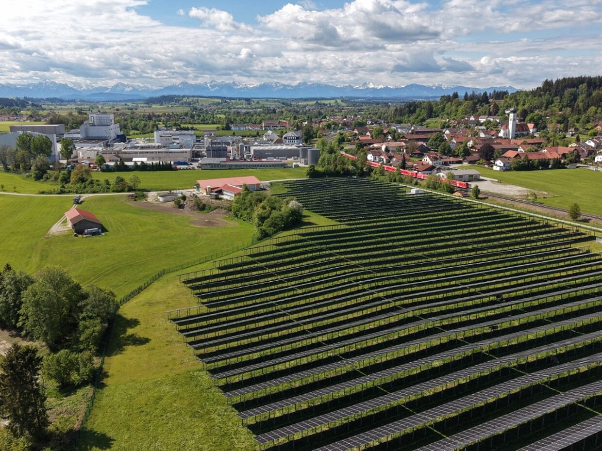 Luftaufnahme eines ländlichen Gebiets mit großen Solaranlagen im Vordergrund, die in Reihen angeordnet sind. Im Hintergrund sieht man eine kleine Ortschaft mit Häusern, einem Wasserreservoir und einer Berglandschaft unter einem teils bewölkten Himmel.
