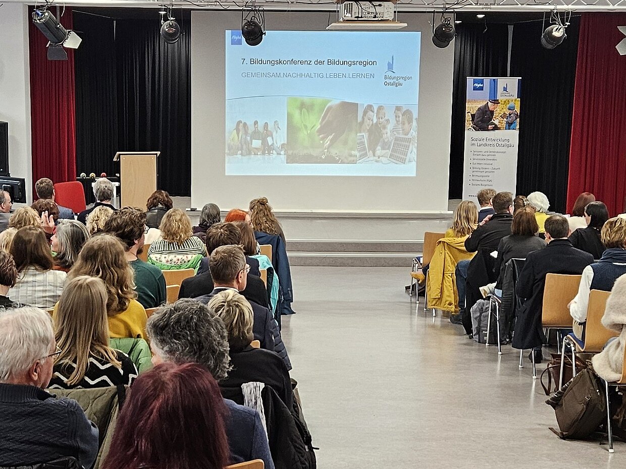 Eine Gruppe von Menschen sitzt in einem Konferenzraum, blickt auf eine präsentiertende Folie mit dem Titel „7. Bildungsakademie der Bildungsregion“. Im Hintergrund sind Stehtische und ein Banner zu sehen, während die Zuschauer an einem Teil der Veranstaltung teilnehmen.