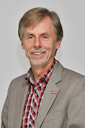 Portrait eines lächelnden Mannes mit blonden Haaren und Bart, der einen grauen Blazer und ein kariertes Hemd trägt. Der Hintergrund ist neutral und hellgrau.