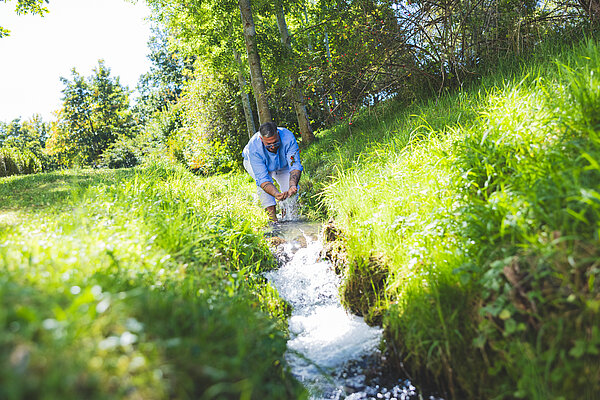 Ein Mann beugt sich über einen kleinen Wasserlauf im Freien, um Wasser zu fangen. Um ihn herum ist saftiges grünes Gras und Bäume. Der Himmel ist blau und sonnig, was eine freundliche und natürliche Umgebung schafft.