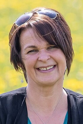 Eine lächelnde Frau mit kurzen, braunen Haaren und Sonnenbrille steht vor einem gelben Hintergrund. Sie trägt eine schwarze Jacke und einen silbernen Anhänger. Die Atmosphäre wirkt freundlich und entspannt.