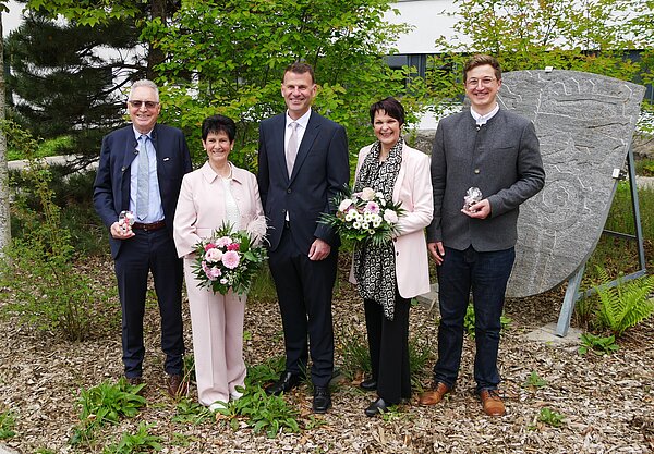 Fünf Personen stehen in einem Außenbereich umgeben von Bäumen und Sträuchern. Sie halten Blumensträuße und posieren freundlich für ein Foto. Im Hintergrund ist ein Stein mit Gravur sichtbar. Die Gruppe scheint an einem festlichen Anlass teilzunehmen.