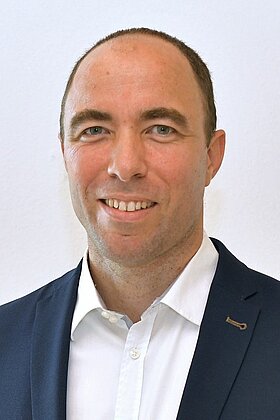 Ein Mann mit kurzem, braunem Haar lächelt in die Kamera. Er trägt ein dunkelblaues Sakko und ein weißes Hemd. Der Hintergrund ist hell und neutral.