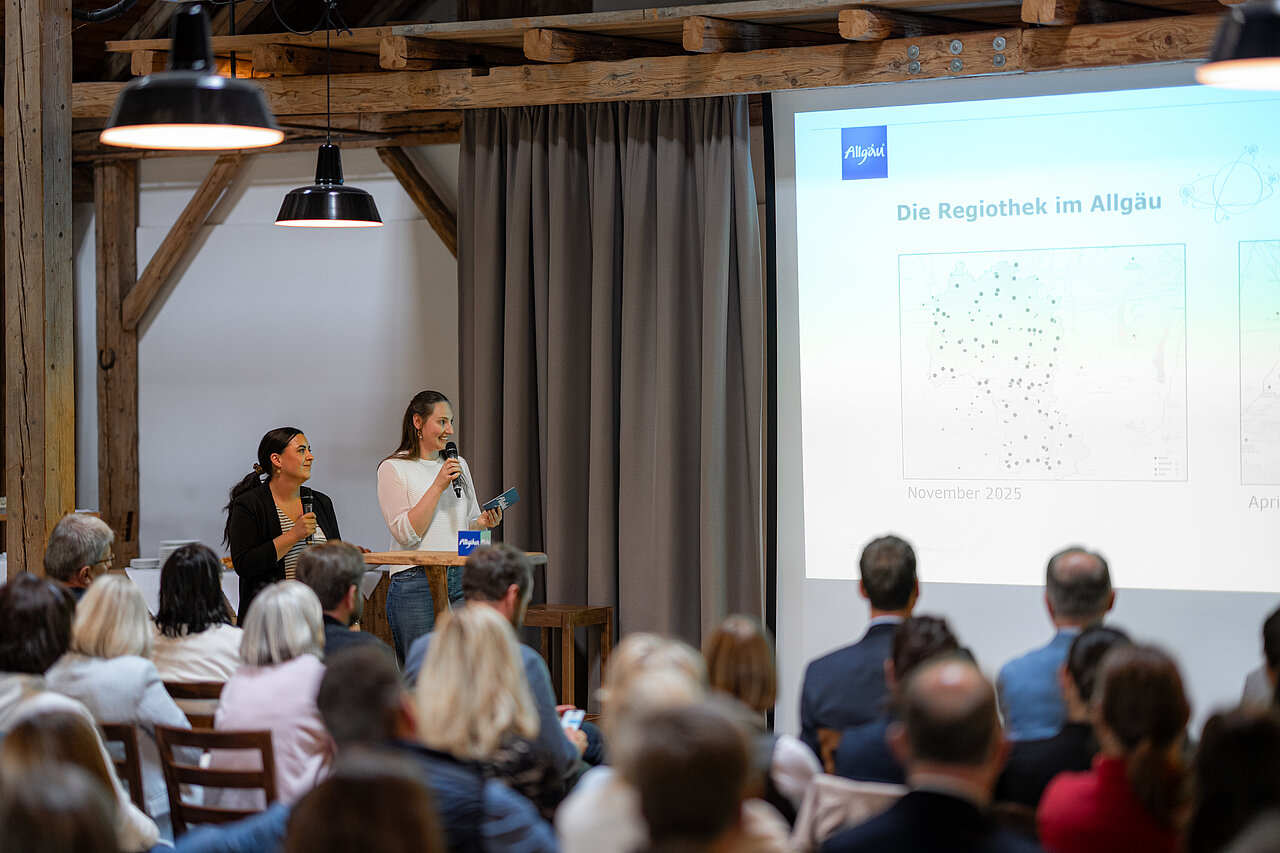 Zwei Frauen präsentieren vor einem Publikum in einem gemütlichen Raum. Eine spricht ins Mikrofon, während die andere Notizen in der Hand hält. Im Hintergrund ist eine Folie mit Karten und dem Titel „Die RegioThek im Allgäu“ zu sehen.