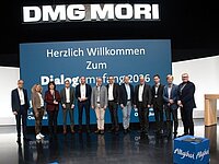 Eine Gruppe von neun Personen steht auf einer Bühne vor einem großen, blauen Banner mit der Aufschrift "Herzlich Willkommen Zum Dialogforum 2016". Im Hintergrund ist das Logo "DMG MORI" zu sehen. Die Personen sind formell gekleidet und lächeln in die Kamera.