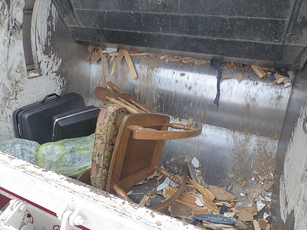 Ein Müllcontainer mit verschiedenen Gegenständen: zwei Koffer, ein gepolsterter Sessel, einige Holzstücke und Abfallbergen. Der Innenraum ist schmutzig und die Wände sind aus Metall.