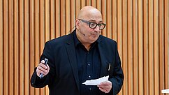 Ein Mann mit Brille und kahlem Kopf steht vor einer Holzverkleidung. Er trägt ein schwarzes Sakko und hält einen Stift in einer Hand sowie ein Blatt Papier in der anderen Hand. Der Mann spricht und wirkt engagiert.
