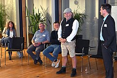 Eine Gruppenansicht von fünf Personen in einem Raum. Eine Frau sitzt auf einem Stuhl und hört zu, während vier Männer stehen oder sitzen, anscheinend an einer Diskussion beteiligt. Im Hintergrund sind Pflanzen und große Fenster sichtbar.