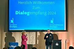 Ein Moderator und eine Moderatorin stehen auf der Bühne vor einer großen Präsentationsleinwand. Auf der Leinwand steht: „Herzlich Willkommen zum Dialogempfang 2024“. Der Raum ist festlich dekoriert, und der Fokus liegt auf den beiden Rednern.