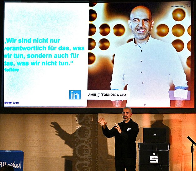 Ein Mann steht auf einer Bühne und spricht. Im Hintergrund ist ein Bildschirm mit einem Zitat und einem Bild von ihm. Das Zitat lautet: „Wir sind nicht nur verantwortlich für das, was wir tun, sondern auch für das, was wir nicht tun.“