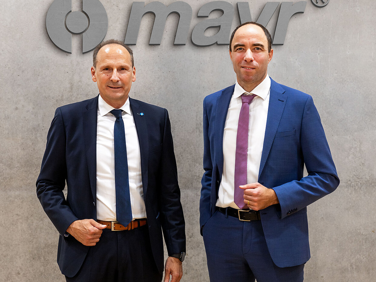 Zwei Männer in Anzügen stehen vor einem grauen Hintergrund mit dem Logo "mayr®". Der erste Mann trägt einen dunkelblauen Anzug und eine Krawatte und hat die Hände in den Taschen. Der zweite Mann trägt einen hellblauen Anzug mit einer lila Krawatte und hält seine Hände vor sich.