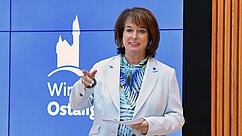 Eine Frau in einem hellen Blazer hält ein Blatt Papier und spricht. Im Hintergrund ist ein blauer Bildschirm mit dem Schriftzug „Windsor Ostallgaus“ und einem Logo, das eine Burg darstellt.