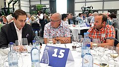 Drei Männer sitzen an einem festlich gedeckten Tisch mit Wasser- und Weingläsern. Sie diskutieren konzentriert. Im Hintergrund sind weitere Personen und Traktoren zu sehen. Eine Tischkarte zeigt die Nummer 15.