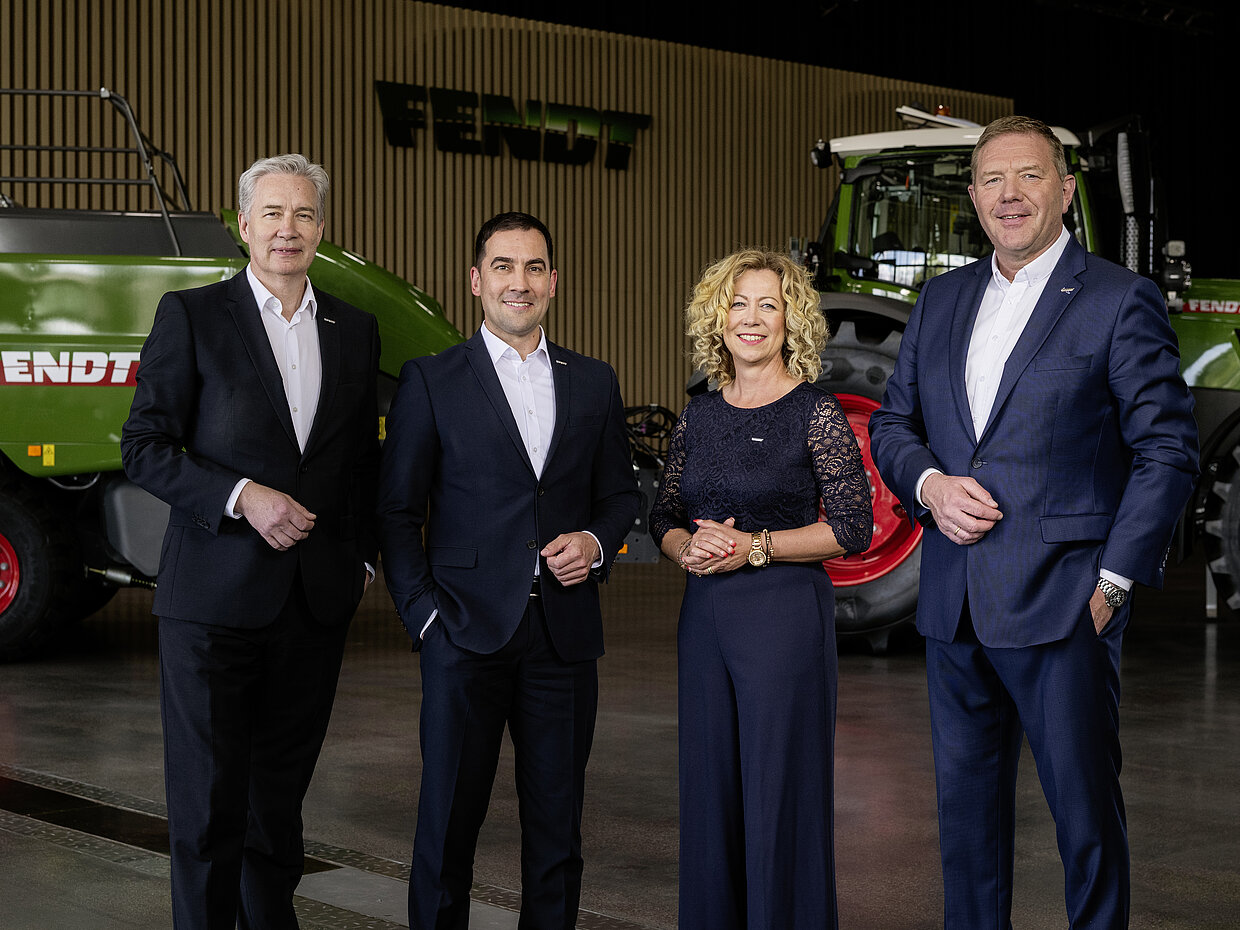 @ AGCO/Fendt Vier Personen stehen in einem modernen Raum, umgeben von landwirtschaftlichen Maschinen. Alle tragen formelle Kleidung und lächeln. Im Hintergrund ist ein großes Fendt-Fahrzeug sichtbar. Das Bild vermittelt ein professionelles und freundliches Ambiente.