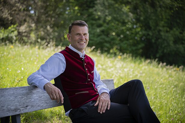 Ein Mann in traditioneller Tracht sitzt entspannt auf einer Bank in einer grünen Landschaft. Er lächelt und trägt ein rotes Wams über einem weißen Hemd. Im Hintergrund sind Bäume und Wiese sichtbar.