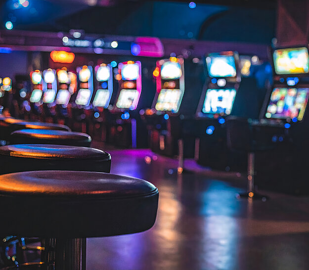 Eine Reihe von Spielautomaten steht in einem beleuchteten Casino. Vor den Automaten sind einige schwarze Barhocker platziert. Die Umgebung ist schwach beleuchtet, mit verschiedenen bunten Lichtern, die eine festliche Atmosphäre schaffen.