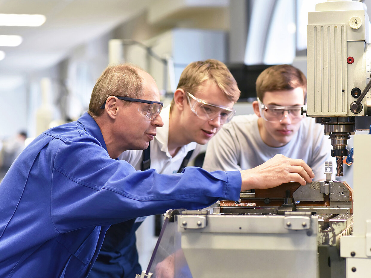 Eine Gruppe von vier Personen in einem technischen Umfeld, darunter ein älterer Mann in Arbeitskleidung und drei jüngere Männer, die Schutzbrillen tragen. Sie beobachten aufmerksam ein Maschinenbedienungsverfahren an einer industriellen Maschine.
