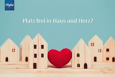 Eine Gruppe von kleinen Holzhäusern steht auf einem Tisch, in der Mitte ein rotes Herz aus Wolle. Im Hintergrund ist ein blauer Himmel sichtbar. Oben links befindet sich ein Logo. Der Text fragt: "Platz frei in Haus und Herz?