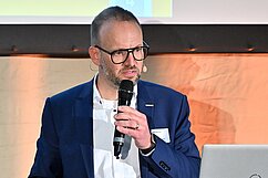 Ein Mann mit Brille trägt einen Anzug und hält ein Mikrofon. Er steht vor einem Projektorbildschirm und spricht. Auf dem Tisch vor ihm steht ein Laptop. Der Hintergrund ist in warmen Tönen beleuchtet.