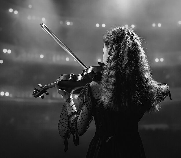 Eine junge Violinistin steht mit dem Rücken zur Kamera auf einer Bühne. Sie trägt ein schwarzes Kleid mit transparenten Ärmeln. Hinter ihr sind verschwommene Lichter eines Publikums zu sehen, die eine besondere Atmosphäre schaffen.