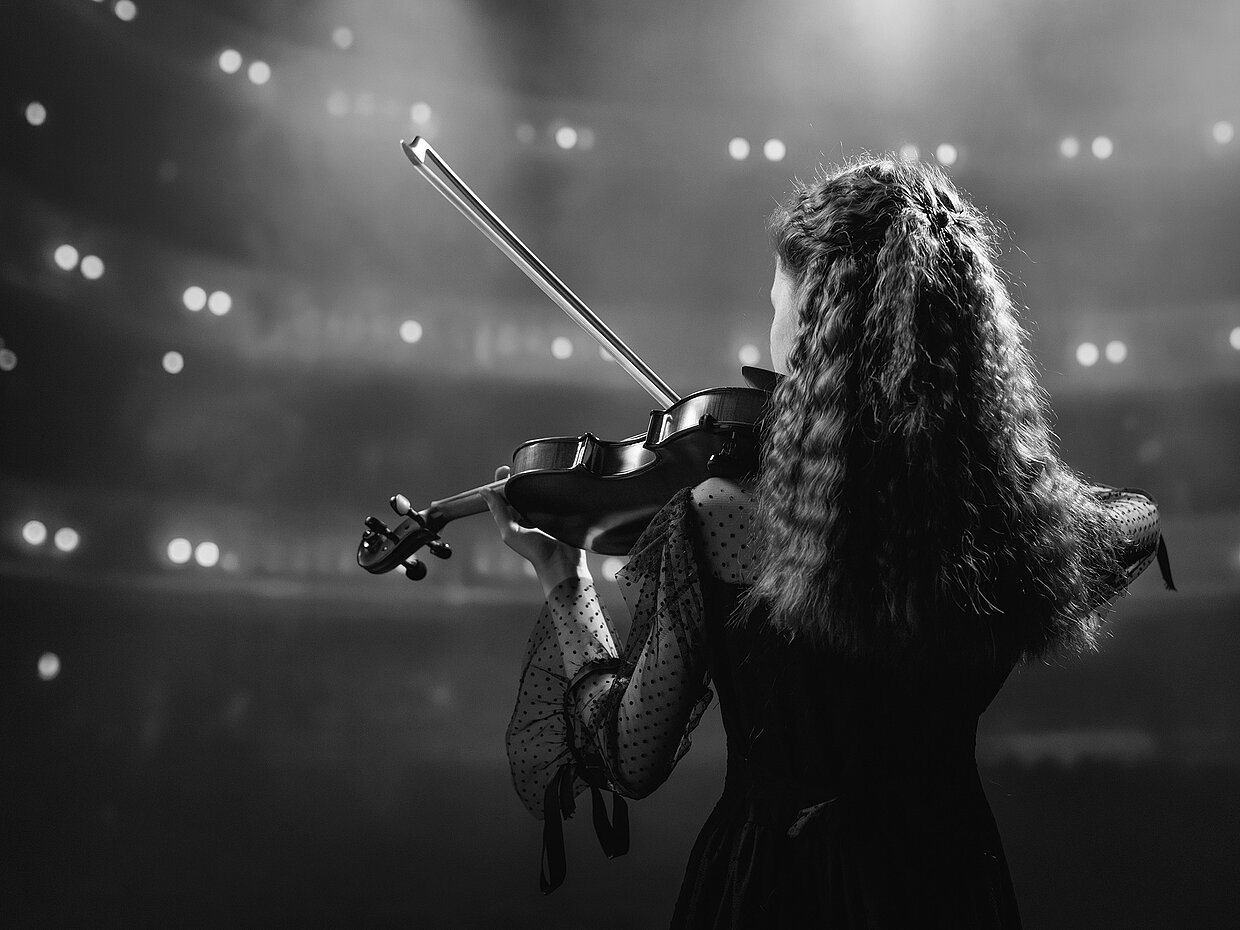 Eine junge Violinistin steht mit dem Rücken zur Kamera auf einer Bühne. Sie trägt ein schwarzes Kleid mit transparenten Ärmeln. Hinter ihr sind verschwommene Lichter eines Publikums zu sehen, die eine besondere Atmosphäre schaffen.