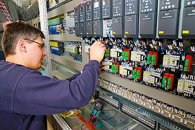 Ein Techniker repariert oder wartet ein Steuerungssystem in einem Schaltschrank. Er konzentriert sich auf die Verdrahtung und den Anschluss von elektrischen Komponenten. Im Hintergrund sind weitere elektronische Geräte und Kabel zu sehen.