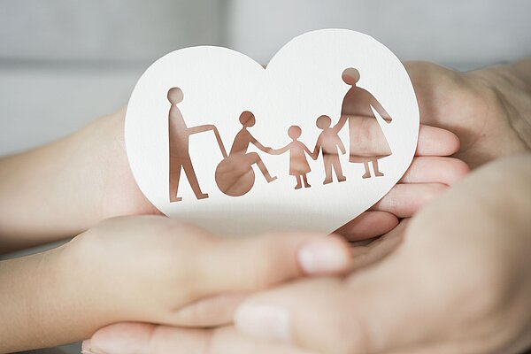 Eine herzförmige Papiergestaltung zeigt eine Familie, die sich an den Händen hält. Im Hintergrund sind zwei Hände zu sehen, die das Herz halten, was Fürsorge und Unterstützung symbolisiert. Die Darstellung umfasst eine Person im Rollstuhl und drei weitere Familienmitglieder.