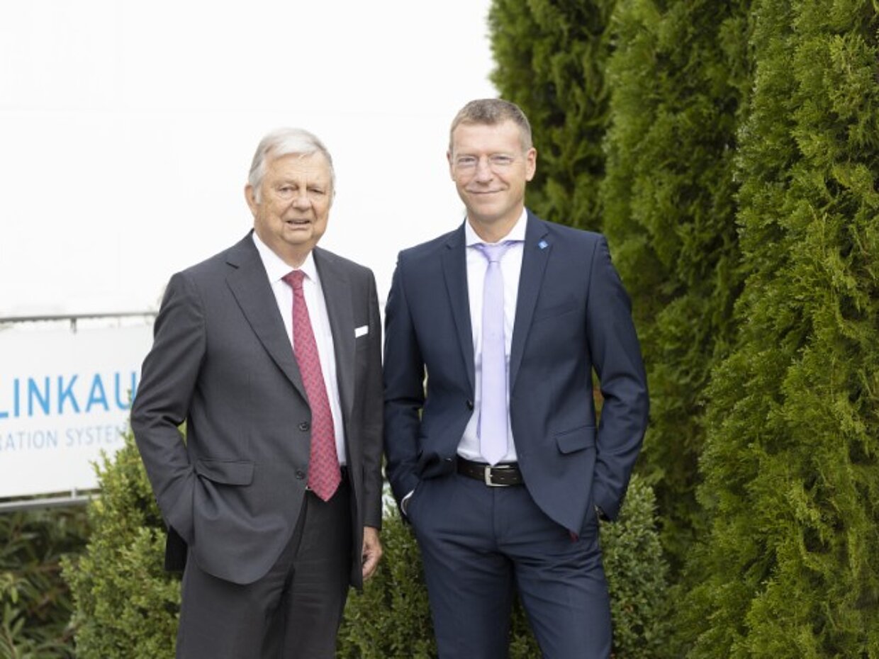 Zwei Männer stehen neben einer Hecke vor einem Schild mit der Aufschrift "Klinkau Filtration Systems". Der ältere Mann trägt einen Anzug mit Krawatte, der jüngere Mann einen dunkelblauen Anzug ohne Krawatte. Beide schauen freundlich in die Kamera.
