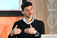 Ein junger Mann mit dunklem Haar und Bart steht an einem Pult und spricht. Er trägt einen schwarzen Pullover über einem weißen Hemd und hat ein Mikrofon am Ohr. Im Hintergrund sind Präsentationsfolien zu sehen.