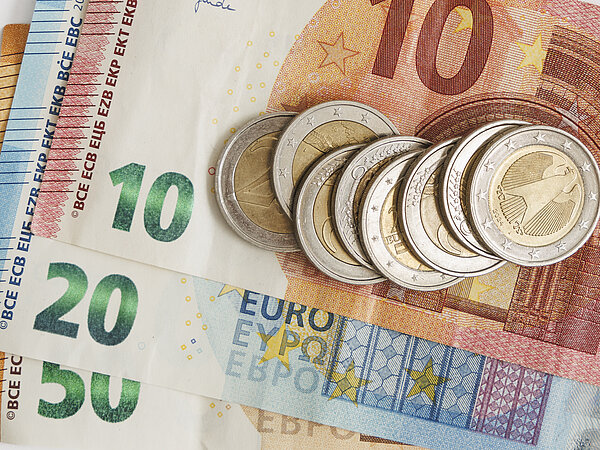 Ein Stapel von Euro-Banknoten in den Werten 10€, 20€ und 50€, mit mehreren 1-Euro-Münzen, die obenauf liegen. Der Hintergrund ist neutral und hebt das Geld hervor.