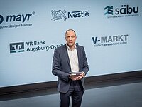 Ein Mann in einem Anzug steht auf einer Bühne vor einem Bildschirm mit Logos von verschiedenen Unternehmen, darunter mayr, Nestlé, säbä, VR Bank Augsburg-Ostalb und V-Markt. Er hält ein Mikrofon oder ein Tablet in der Hand.