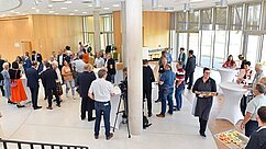 Eine Gruppe von Menschen in einem modernen Veranstaltungsraum, die sich bei einem Networking-Event versammeln. Einige Personen sprechen miteinander, während andere an Stehtischen stehen oder Essen servieren. Helle Holz- und Glasarchitektur prägt die Umgebung.