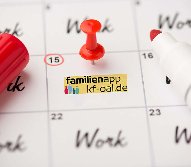 Ein Kalenderblatt mit dem Datum 15, markiert mit einer roten Pinnnadel. Daneben liegt ein roter Marker. Auf dem Kalender steht "Work" und das Logo einer Familien-App mit der URL "kf-oal.de".