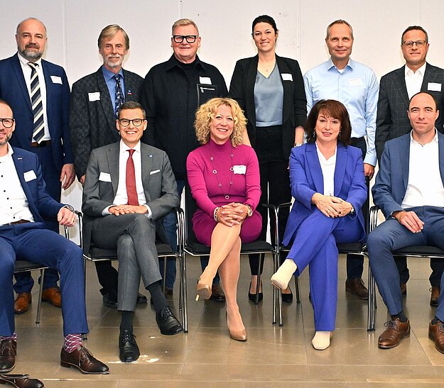 Eine Gruppe von 14 Personen steht und sitzt in zwei Reihen. Sie tragen formelle Kleidung und lächeln. Im Hintergrund ist eine neutrale Wand sichtbar. Jeder hat ein Namensschild an der Kleidung, das auf eine geschäftliche Veranstaltung hindeutet.