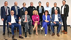 Eine Gruppe von 14 Personen steht und sitzt in zwei Reihen. Sie tragen formelle Kleidung und lächeln. Im Hintergrund ist eine neutrale Wand sichtbar. Jeder hat ein Namensschild an der Kleidung, das auf eine geschäftliche Veranstaltung hindeutet.