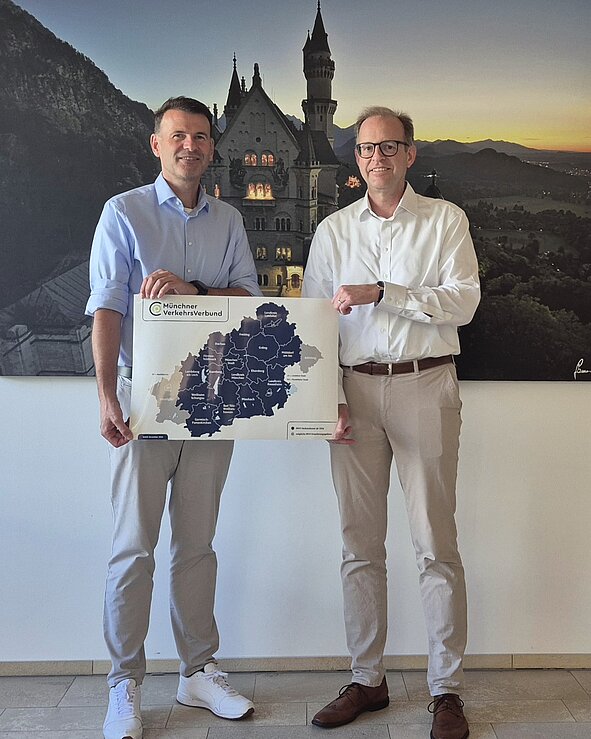 Zwei Männer stehen in einem Büro und halten eine Karte des "Werkverbund". Im Hintergrund ist ein Bild eines Schlosses in einer bergigen Landschaft zu sehen. Beide tragen legere Kleidung.