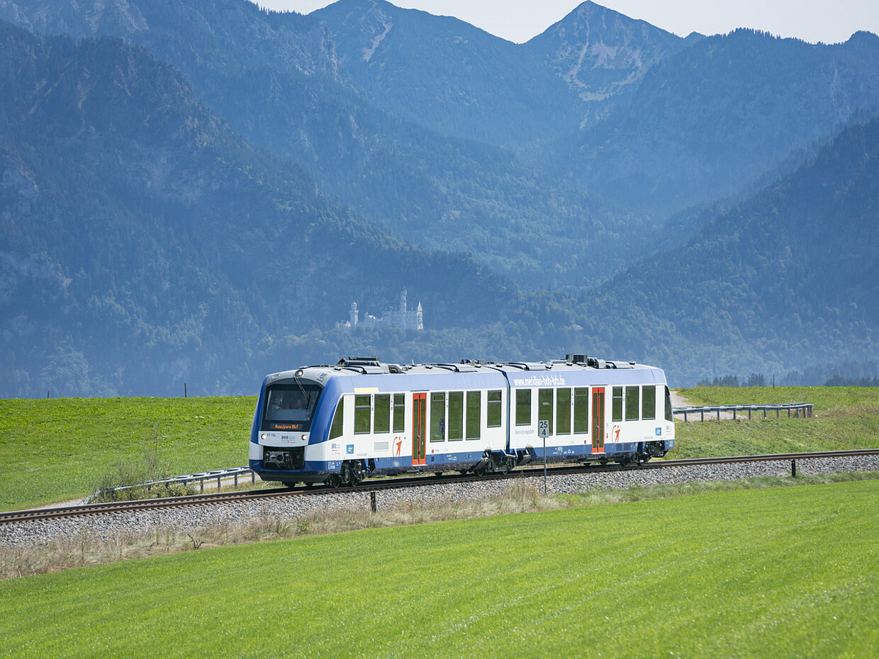 Ein Zug fährt auf einer Gleisanlage durch eine grüne Wiesenlandschaft, umgeben von Bergen im Hintergrund. Die Szene zeigt eine klare, sonnige Tagesstimmung. In der Ferne ist ein Schloss sichtbar.