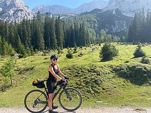 Eine Person steht neben einem Fahrrad auf einem Waldweg. Die Umgebung zeigt grüne Wiesen und hohe Bäume, mit Bergen im Hintergrund. Die Person trägt eine schwarze Radbekleidung und einen Helm, und sie lächelt.